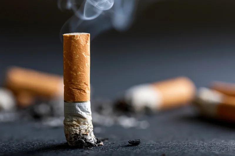 A smoking cigarette butt standing upright on a dark textured surface with smoke rising and ash scattered around