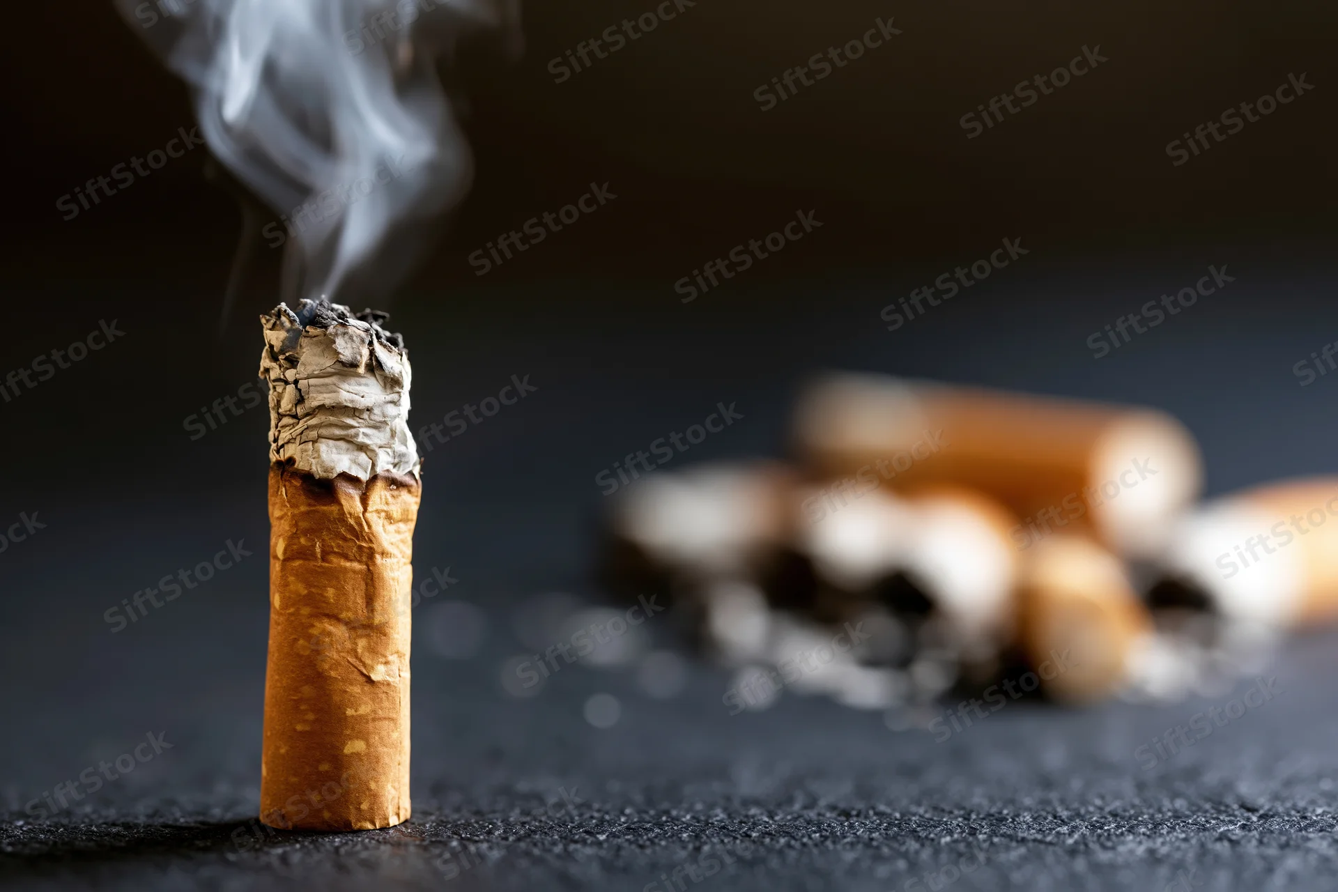 Burning cigarette butt with smoke rising on dark surface, surrounded by discarded butts