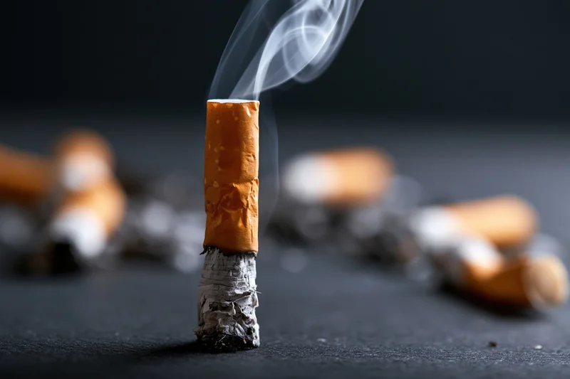A smoking cigarette butt standing upright with smoke rising from it on a dark surface surrounded by other discarded butts