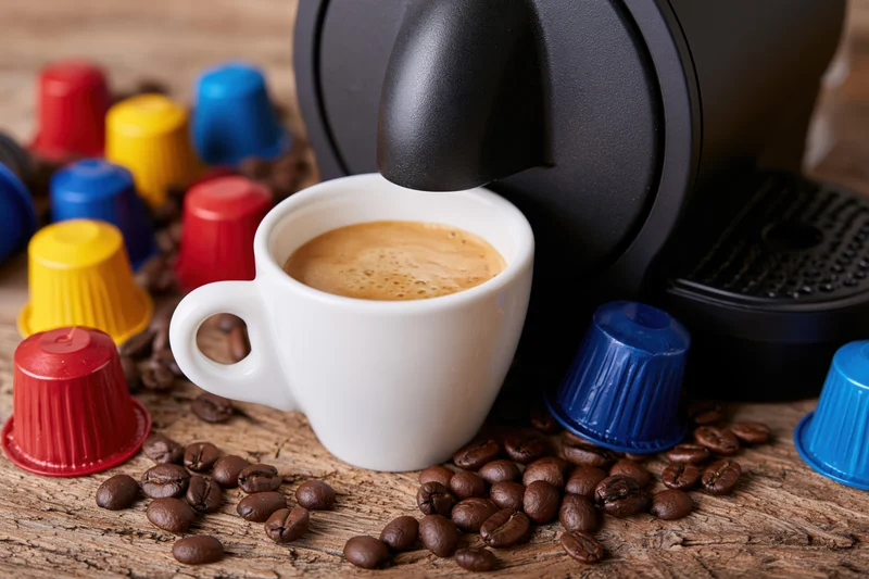 Coffee machine brewing espresso into a white ceramic cup on a wooden table surrounded by coffee beans and colorful capsules