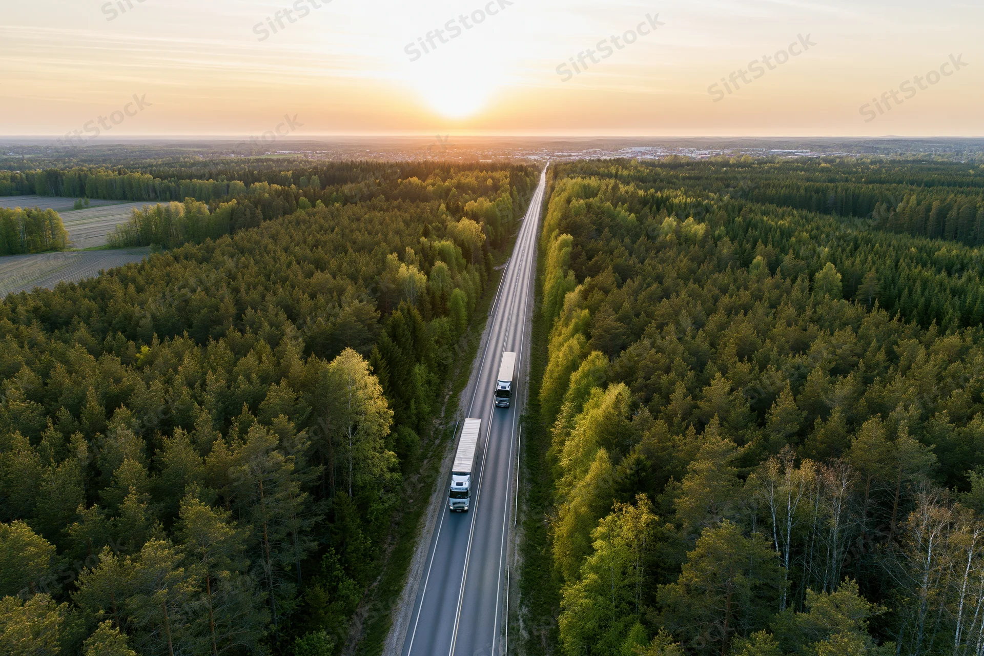 Aerial view of trucks driving on a straight highway through dense forest at sunset