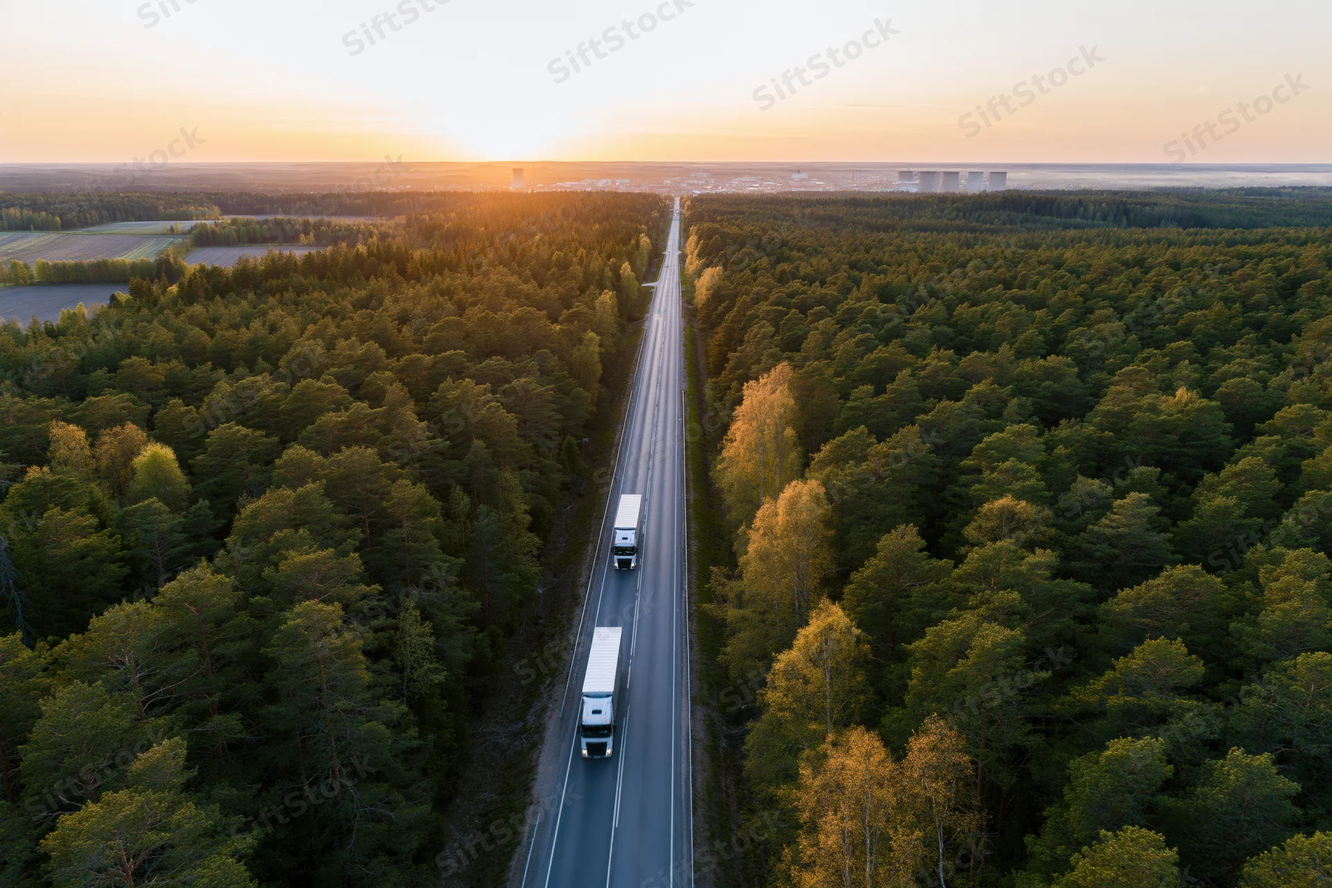Aerial view of two trucks driving on a straight two-lane highway through a dense forest at sunset