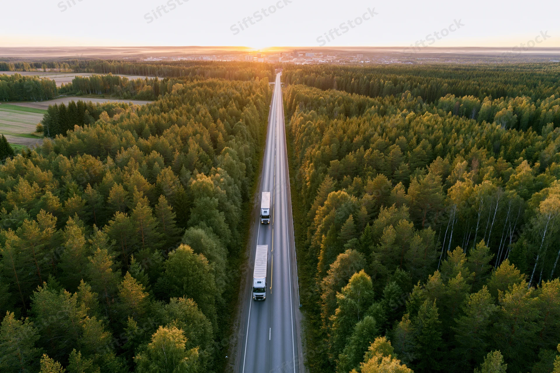 Aerial view of two trucks driving on a straight highway through dense forest at sunset