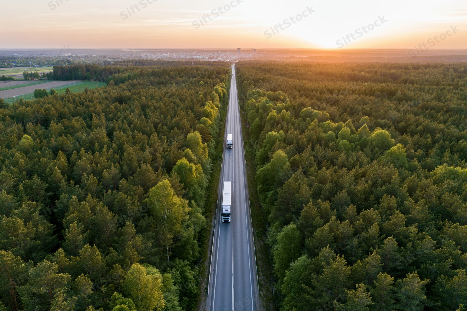 Aerial view of trucks driving on a straight highway through dense forest at sunset