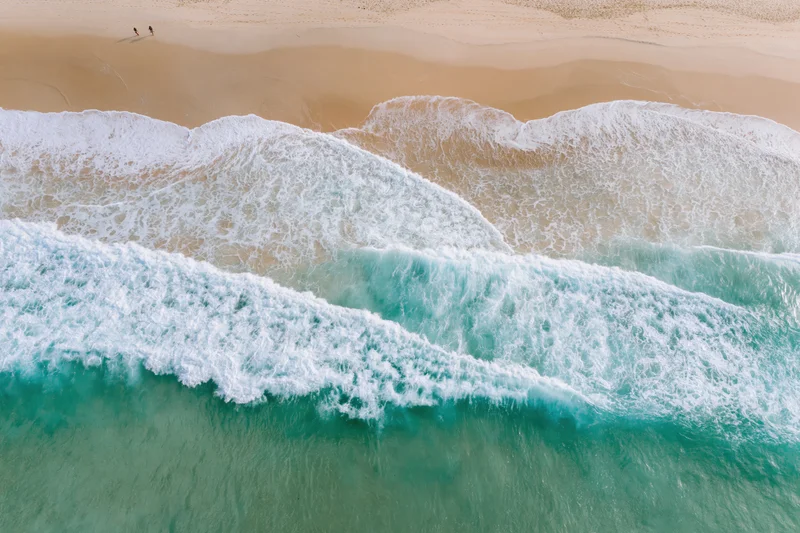 Aerial view of waves breaking on a sandy beach with two people walking along the shore