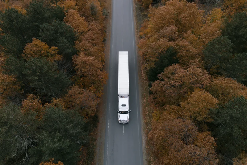 White semi-truck driving down a straight road through autumn forest from overhead drone view