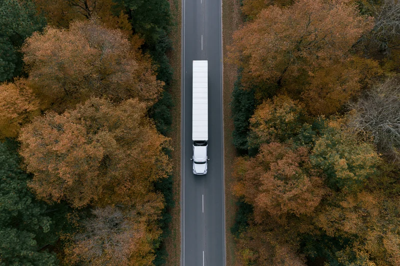 White semi-truck driving on a straight road through an autumn forest from above