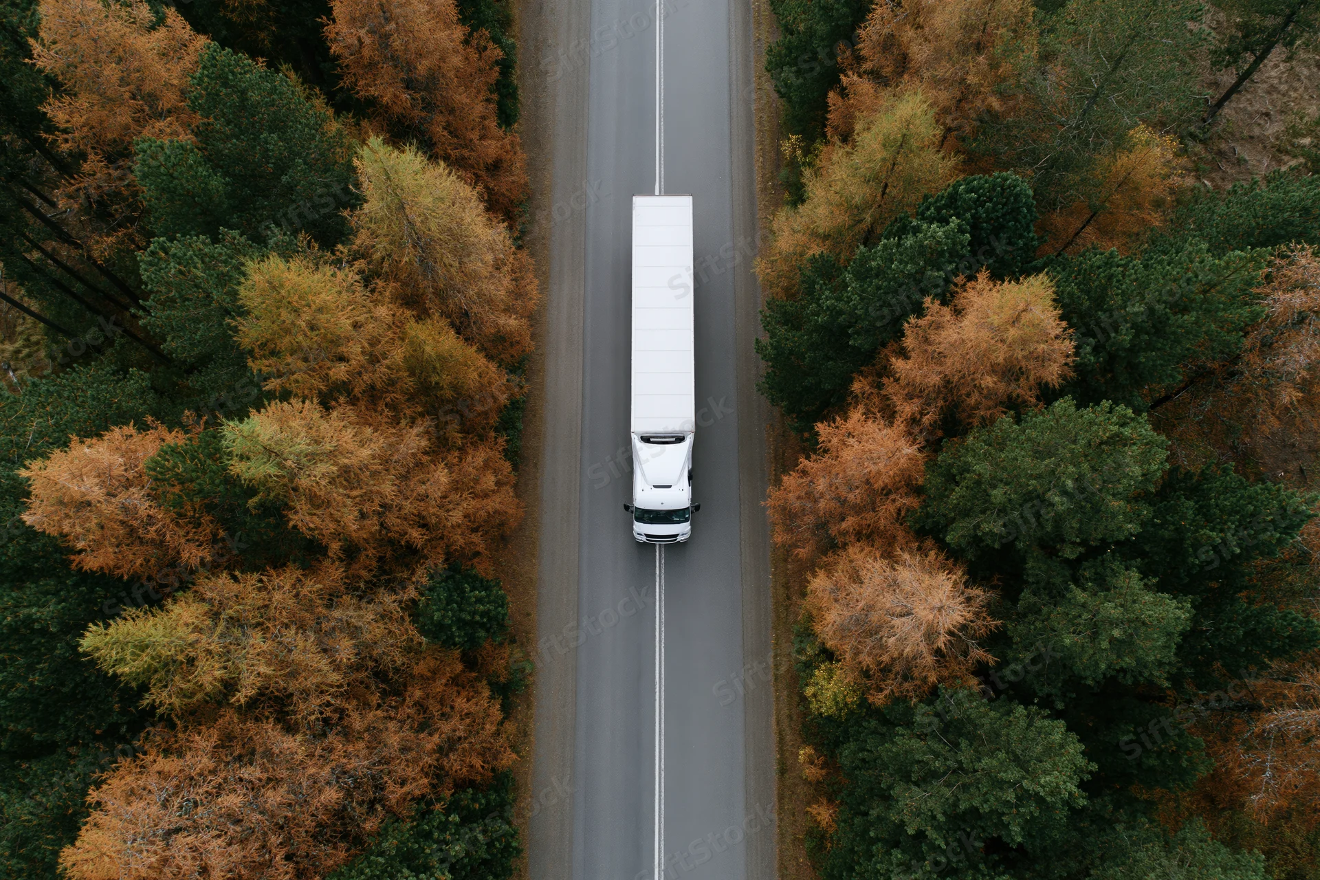 White semi-truck driving on a straight road through an autumn forest viewed from above