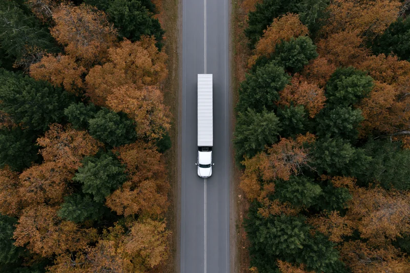 White semi-truck driving on a straight road through autumn forest from overhead drone view