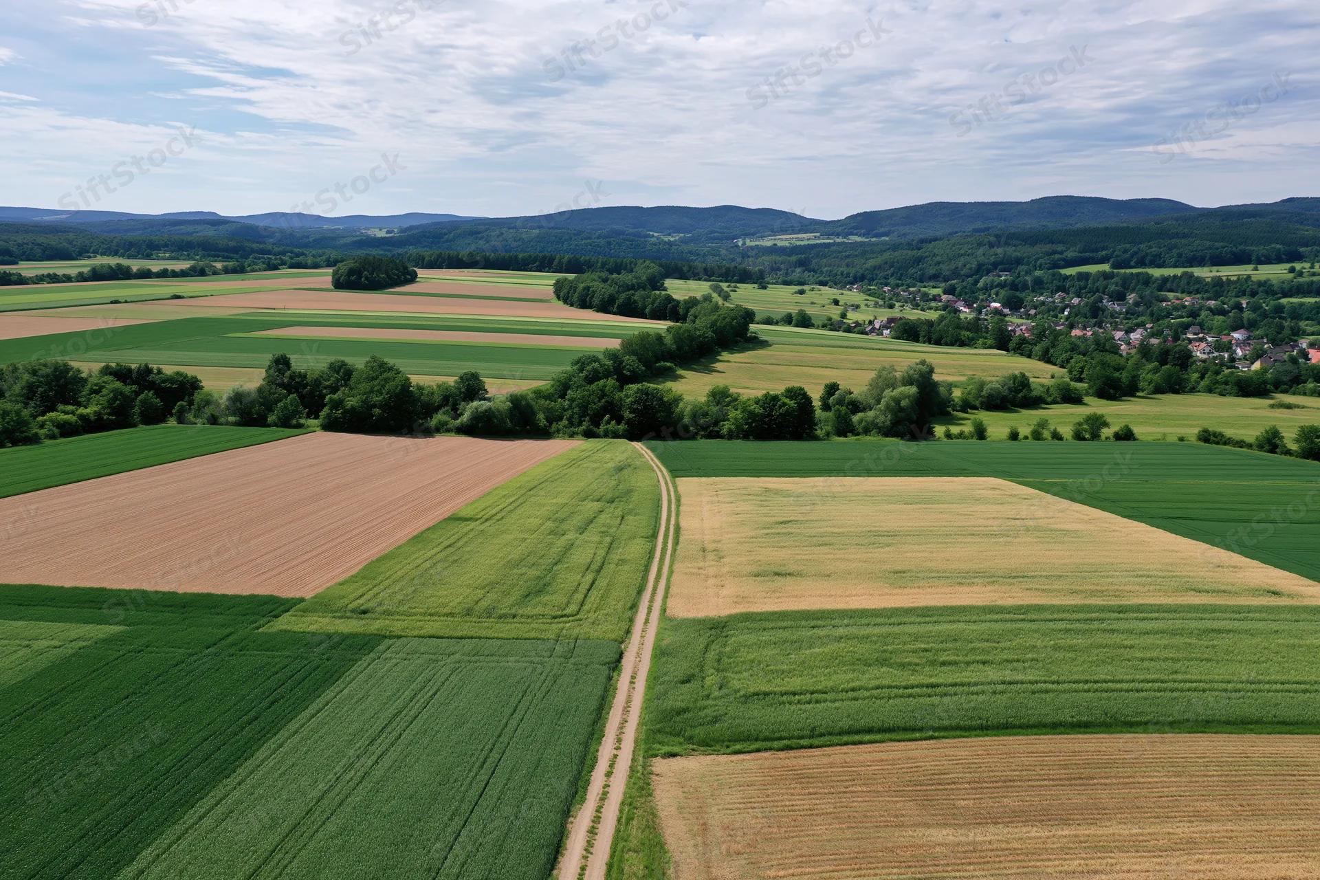 Aerial drone view of patchwork farmland with green and brown fields, trees, and distant village under partly cloudy sky