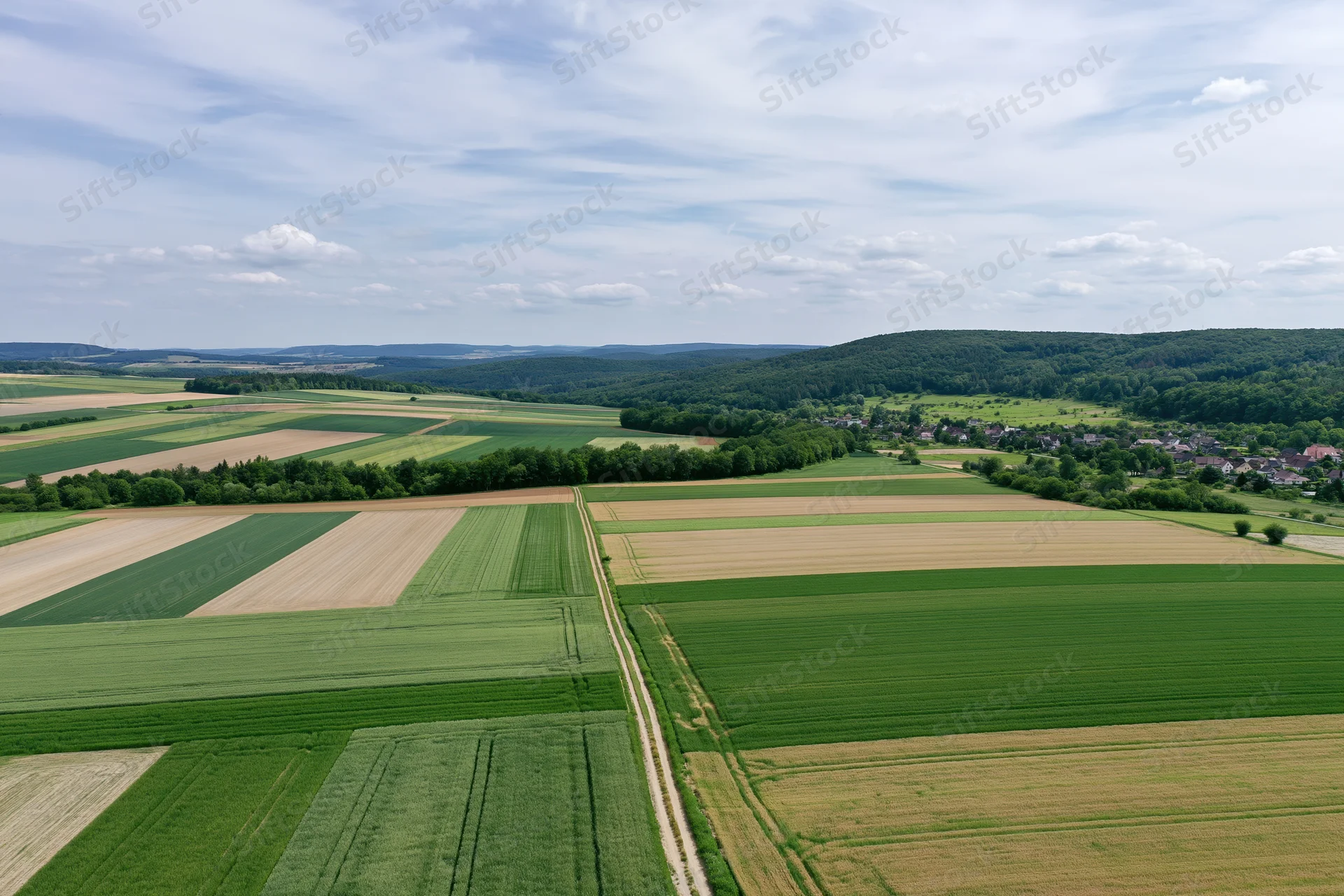 Aerial drone view of patchwork farmland with green and brown fields, trees, and a village in the distance under a partly cloudy sky