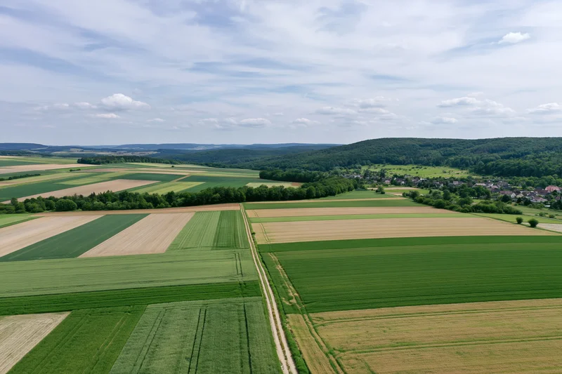 Aerial drone view of patchwork farmland with green and brown fields, trees, and a village in the distance under a partly cloudy sky