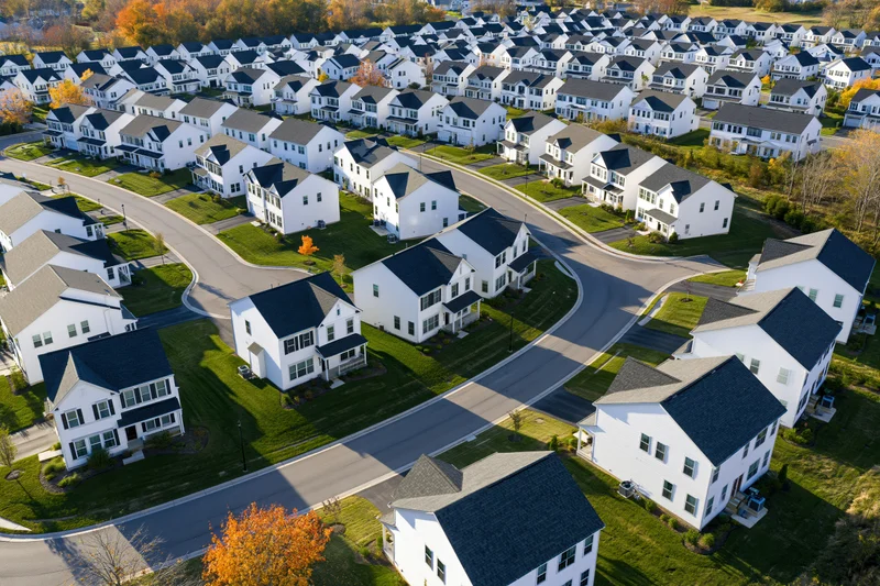 Aerial drone view of a new suburban housing development with white houses and winding streets surrounded by autumn trees