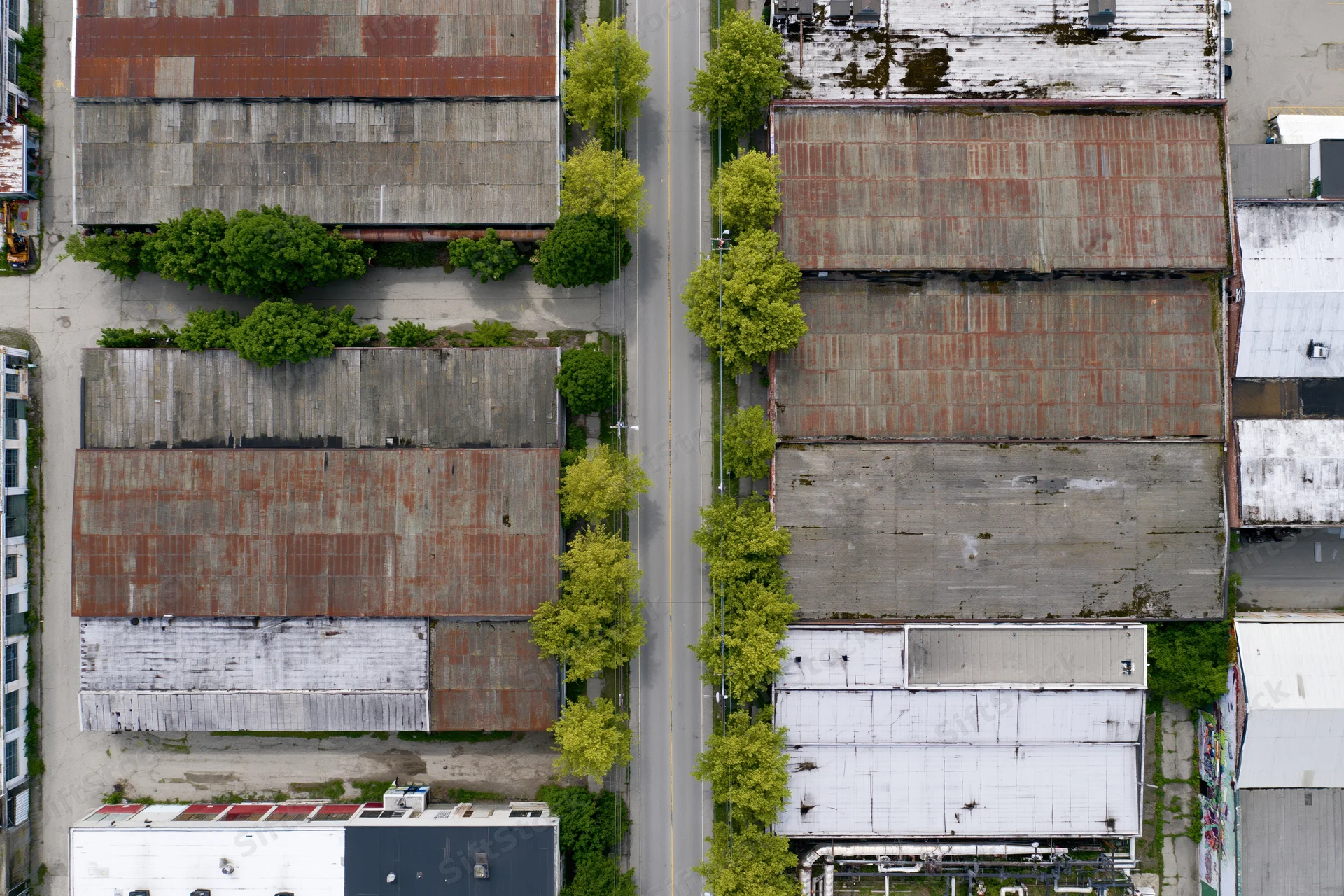 Aerial top-down view of aging industrial buildings with rusted metal roofs and green trees along a street