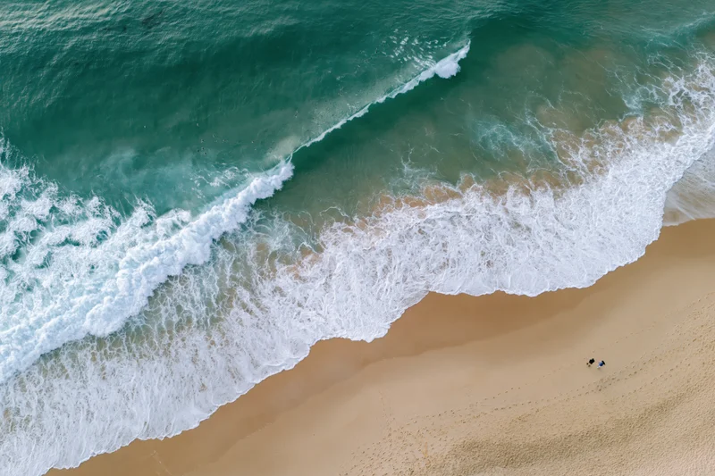 Aerial top-down view of ocean waves breaking on sandy beach with two people walking along shore