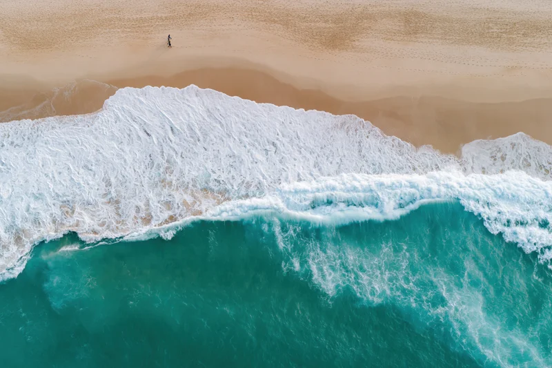Aerial top-down view of turquoise ocean waves breaking on sandy beach with solitary person walking in distance