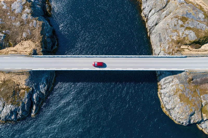 Red car driving across narrow bridge spanning dark blue water between rocky cliffs, aerial top-down view