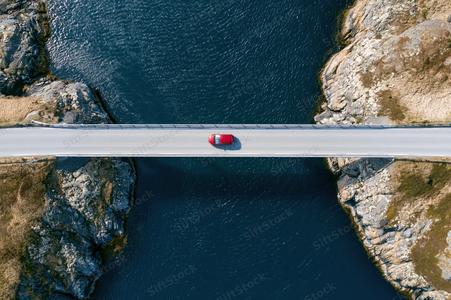 Red car driving across narrow bridge over deep blue river flanked by rocky cliffs, aerial top-down view