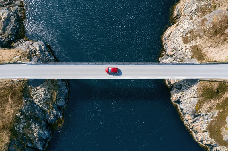 Red car driving across narrow bridge over deep blue river flanked by rocky cliffs, aerial top-down view