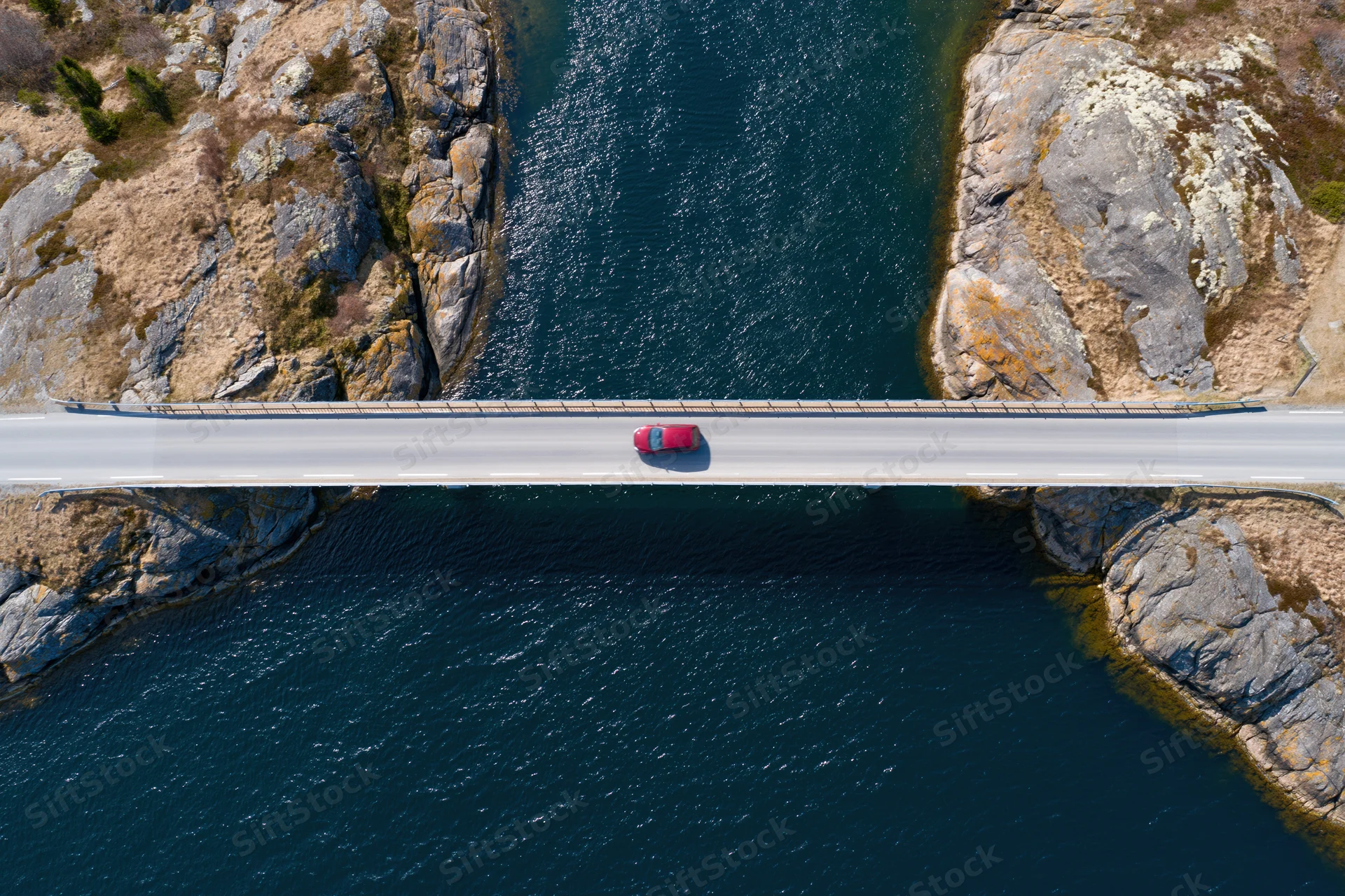 Red car driving across narrow bridge spanning deep blue river between rocky cliffs, aerial top-down view