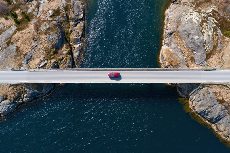 Red car driving across narrow bridge spanning deep blue river between rocky cliffs, aerial top-down view