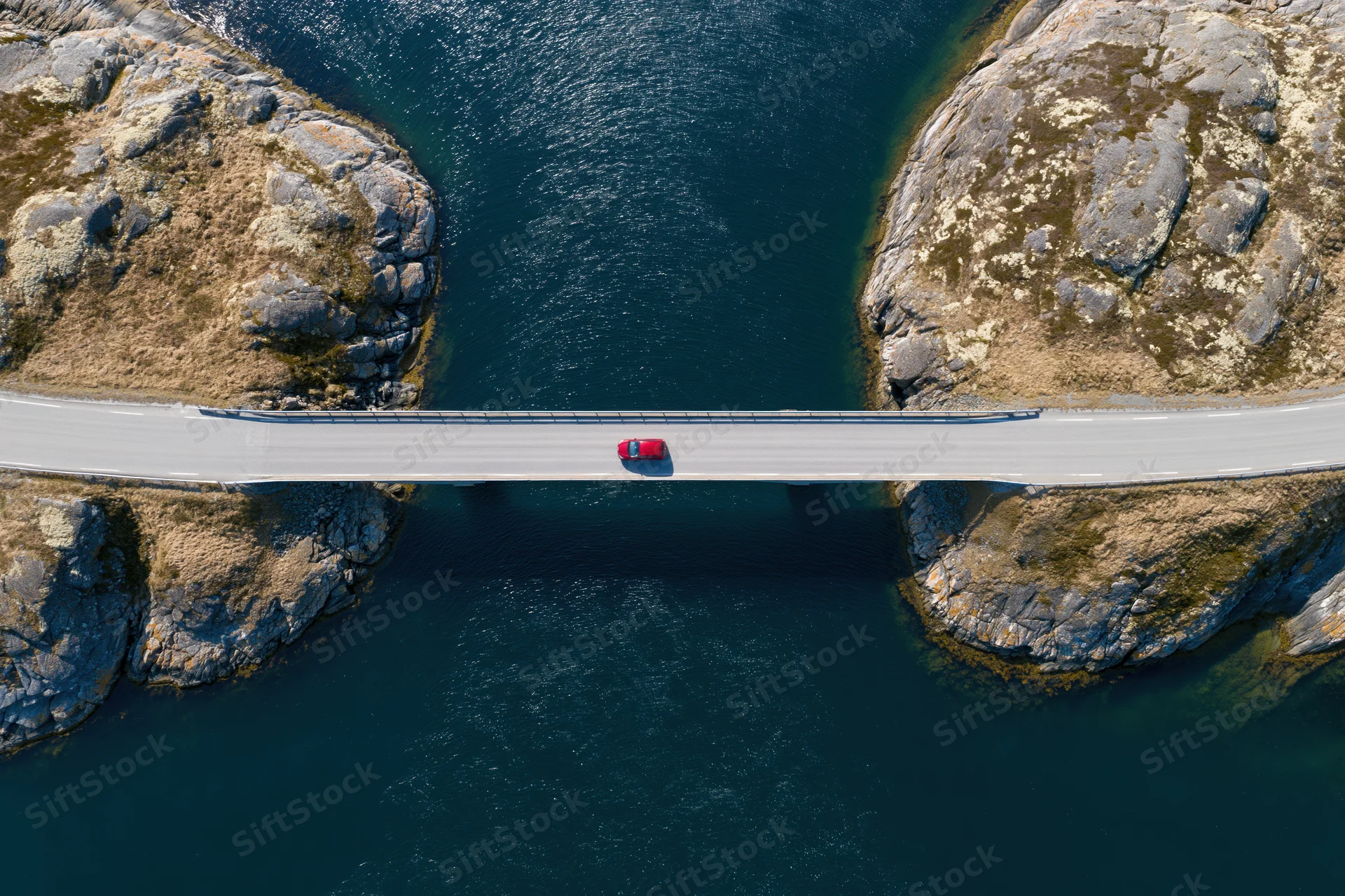 Red car driving across narrow bridge spanning deep blue water between rocky cliffs, aerial top-down view