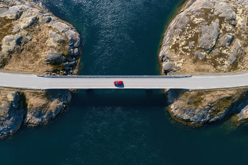 Red car driving across narrow bridge spanning deep blue water between rocky cliffs, aerial top-down view