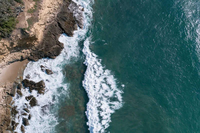 Aerial top-down view of waves crashing against rocky cliff edge along coastline