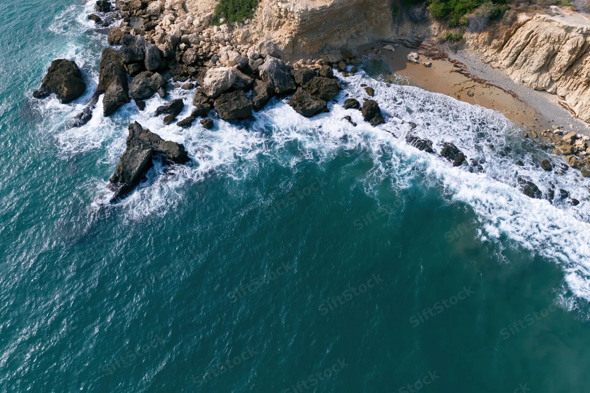 Aerial top-down view of waves crashing against rocky cliff edge meeting sandy shore