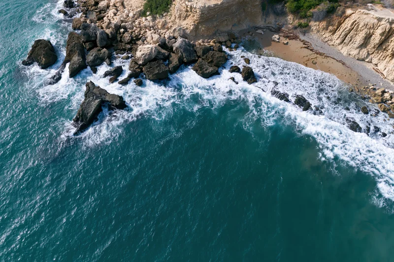 Aerial top-down view of waves crashing against rocky cliff edge meeting sandy shore