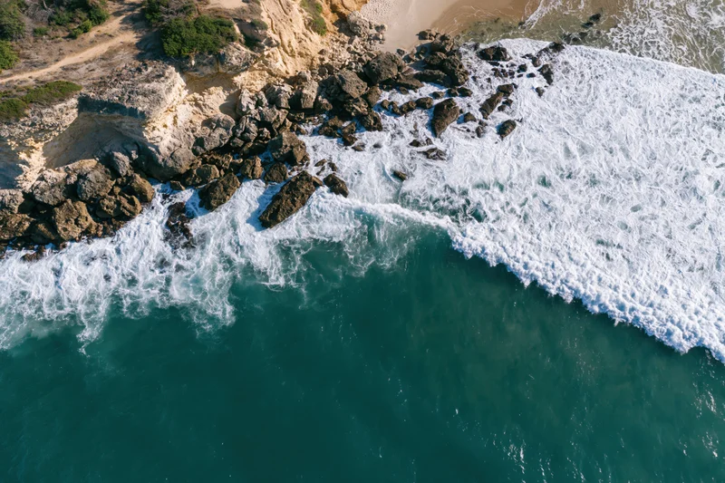 Aerial top-down view of waves breaking against rocky cliff edge meeting sandy beach