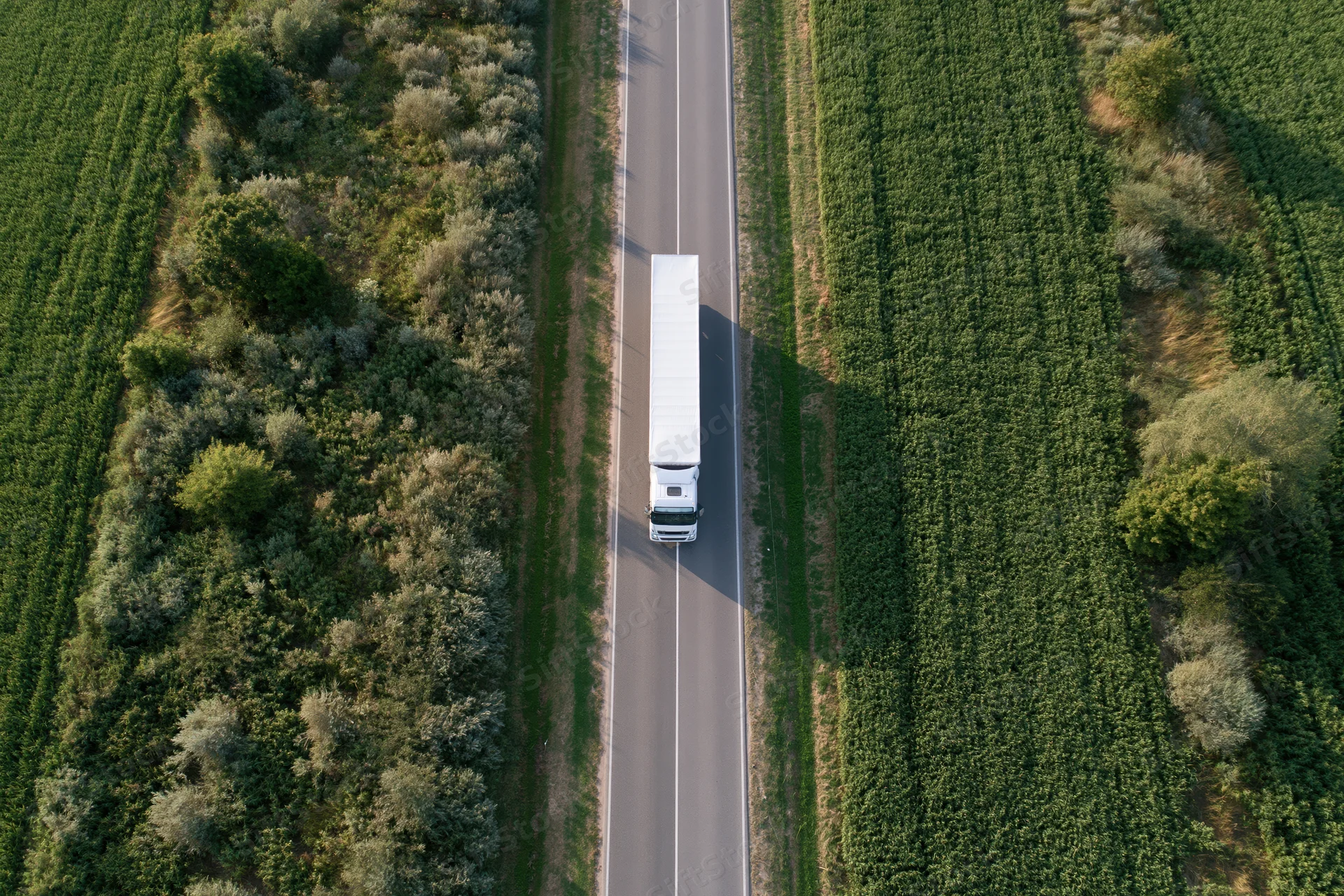 White semi truck driving on a straight highway through green farmland fields