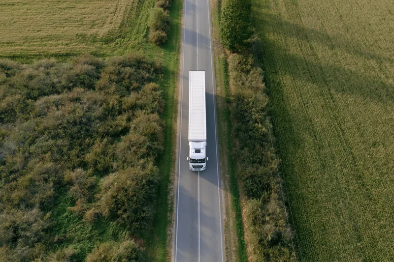 White semi truck driving on a rural highway surrounded by green fields and farmland