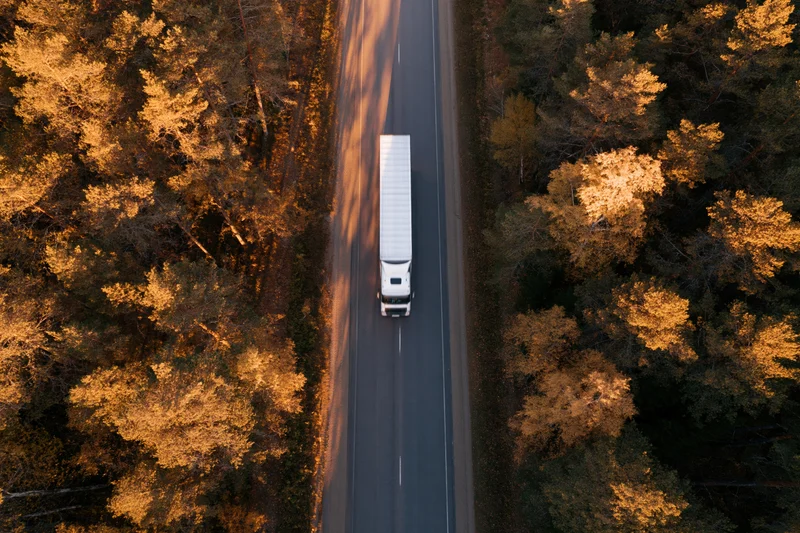 White semi-truck with trailer driving on highway through autumn forest from aerial top-down view