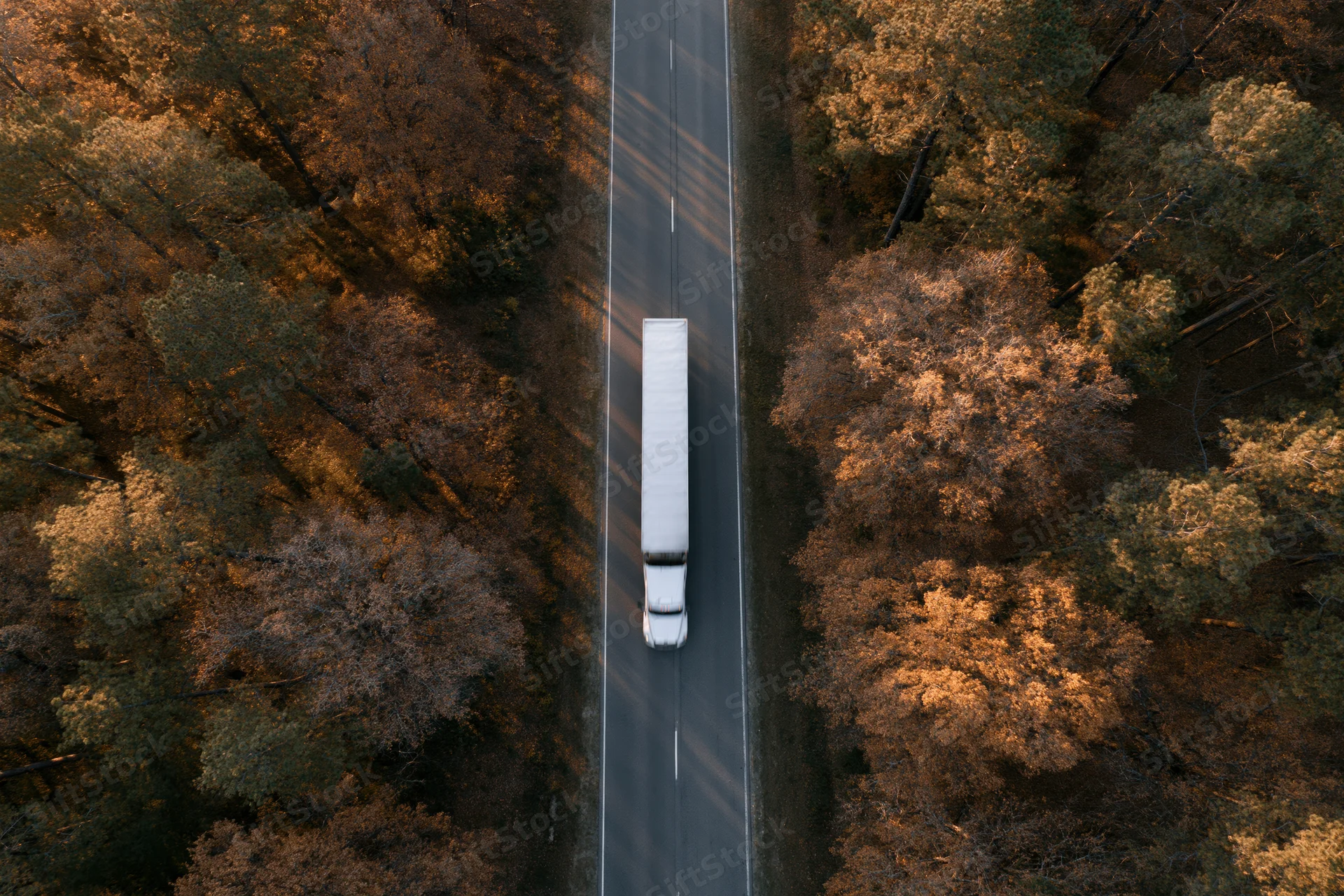 White semi-truck with trailer driving on a straight road through autumn forest from aerial top-down view
