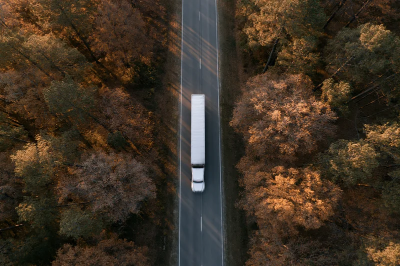 White semi-truck with trailer driving on a straight road through autumn forest from aerial top-down view