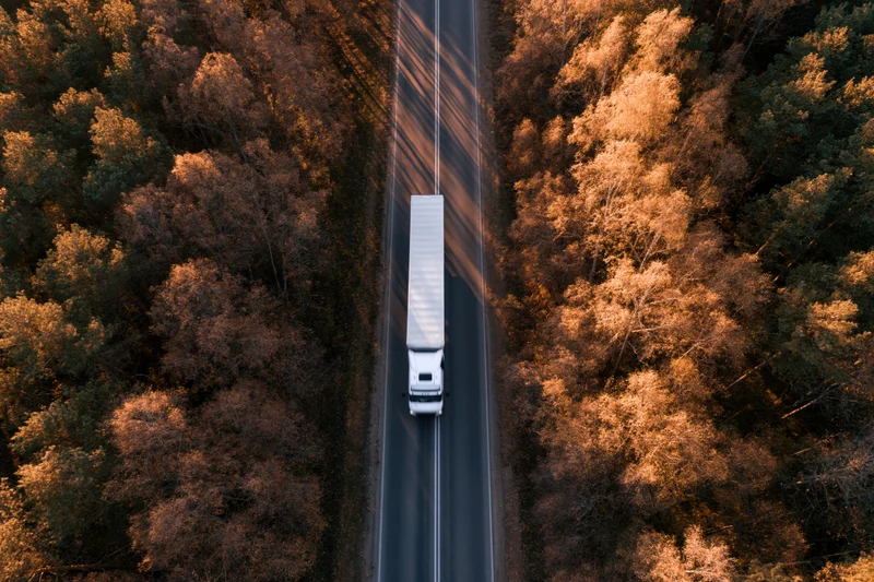 White semi-truck with trailer driving on a straight highway through autumn forest from aerial top-down view