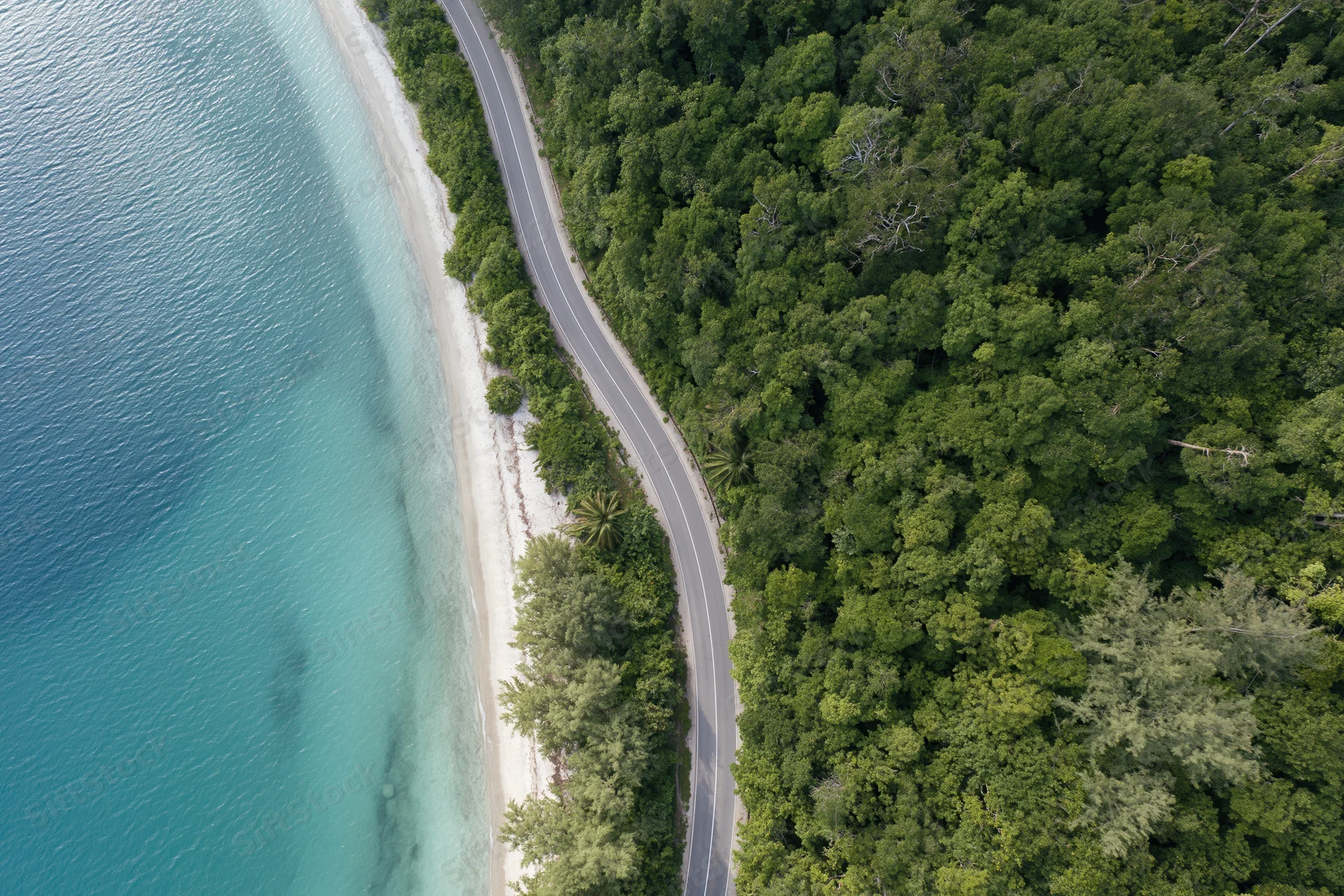 Aerial top-down view of a winding coastal road alongside a white sandy beach and turquoise ocean bordered by dense green forest