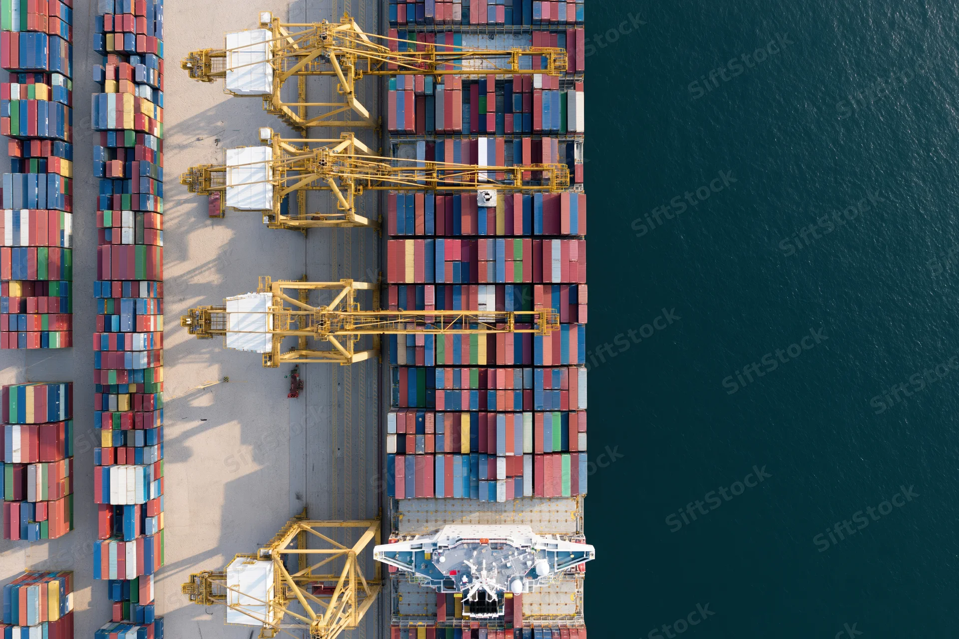 Aerial top-down view of a busy commercial shipping port with stacked containers, cranes, and a cargo ship docked at the terminal