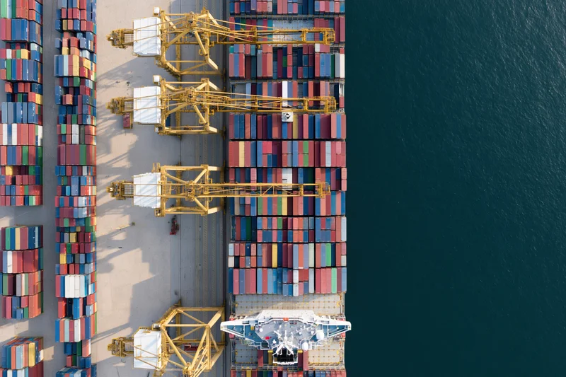 Aerial top-down view of a busy commercial shipping port with stacked containers, cranes, and a cargo ship docked at the terminal