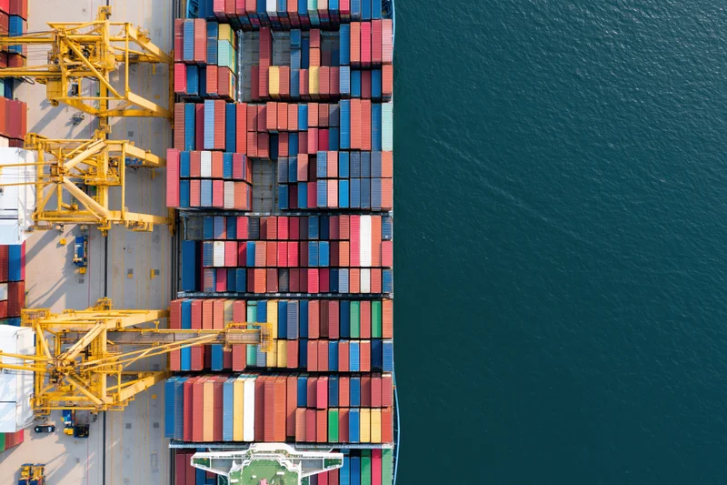 Aerial top-down view of a container ship docked at a busy port with yellow cranes and stacked cargo containers alongside the water