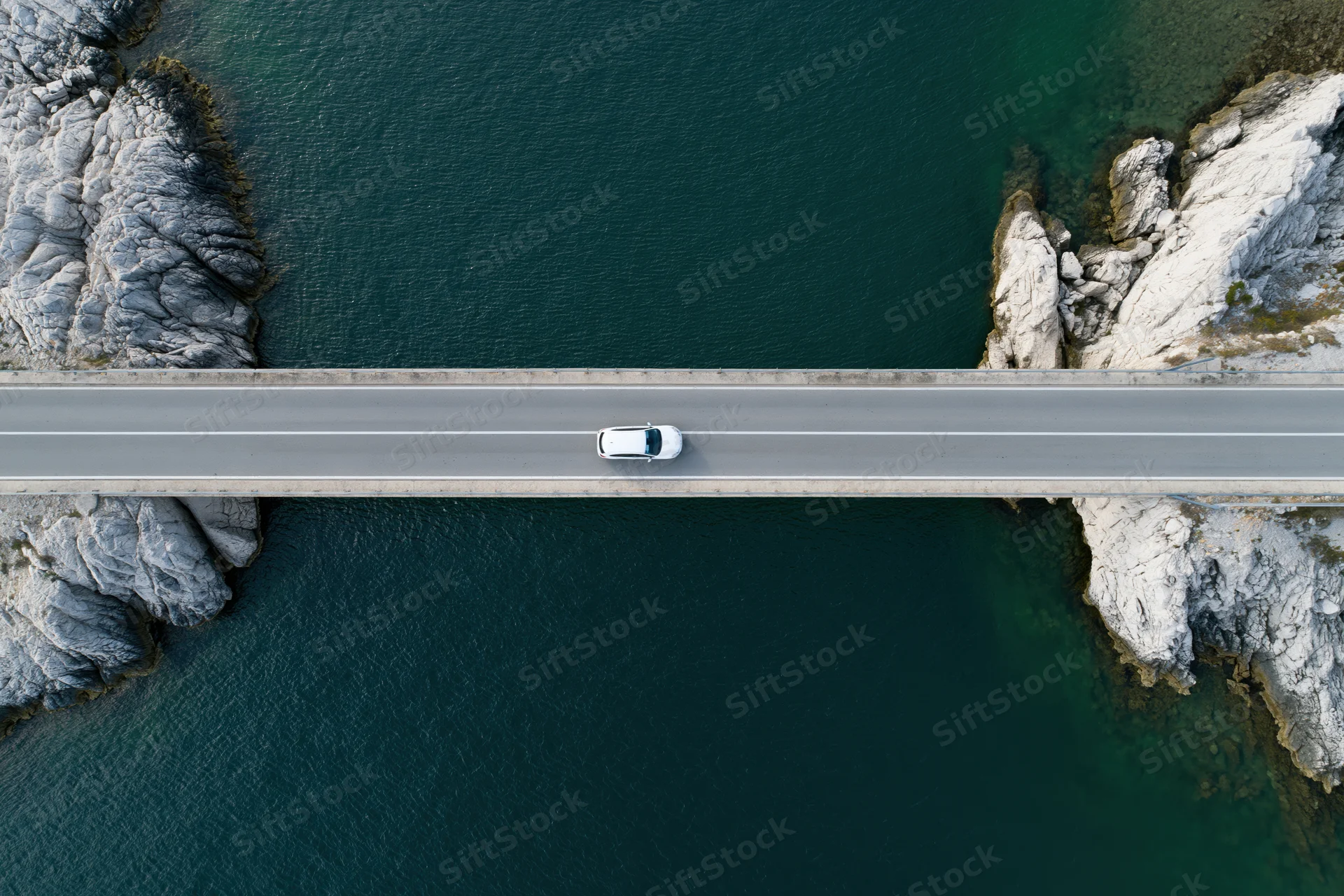 White car driving across narrow concrete bridge over turquoise water between rocky cliffs, aerial top-down view