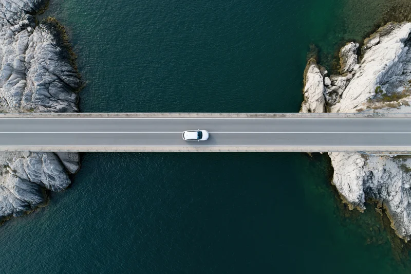 White car driving across narrow concrete bridge over turquoise water between rocky cliffs, aerial top-down view