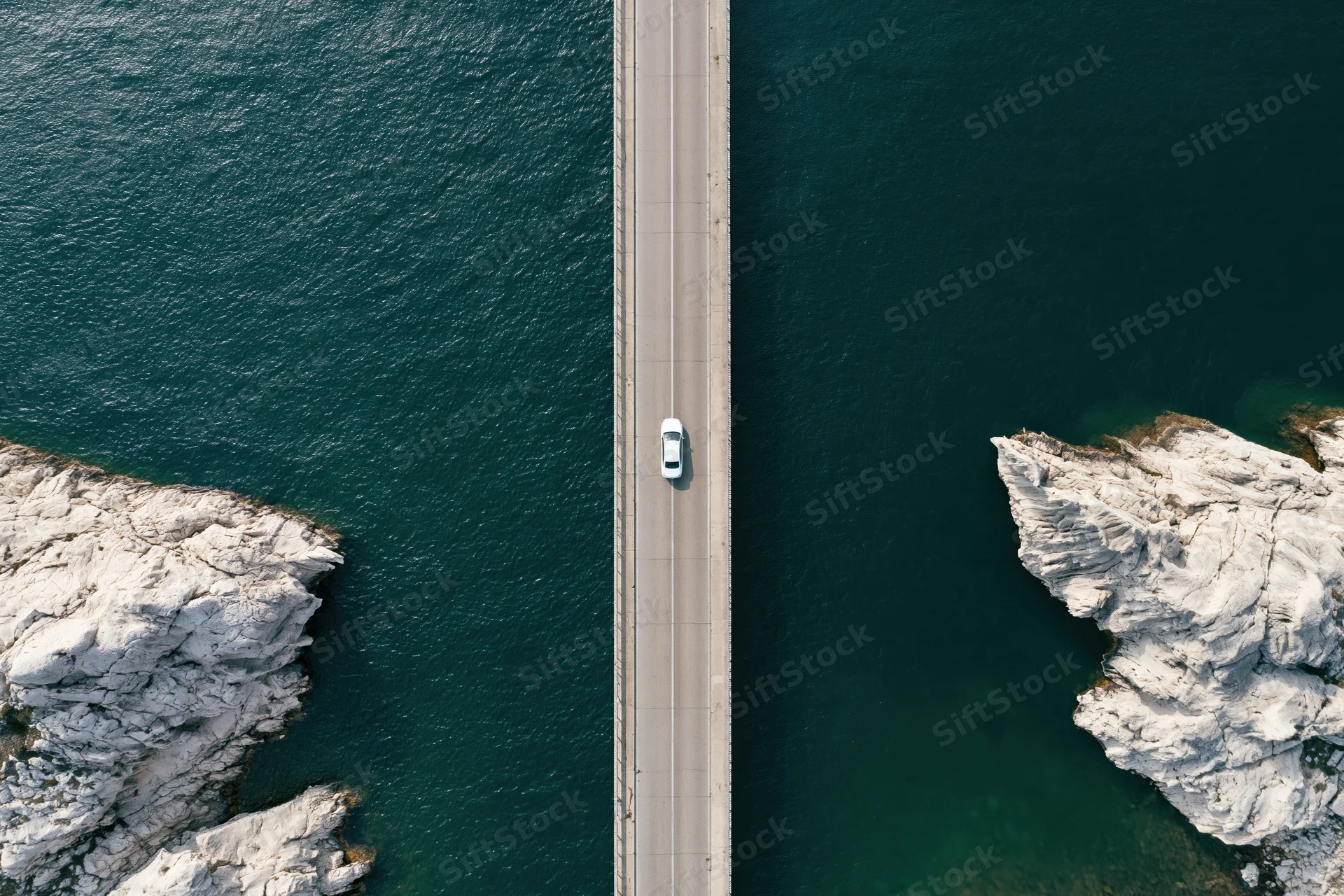 Aerial top-down view of a white car driving on a narrow concrete bridge over deep blue water between rocky cliffs