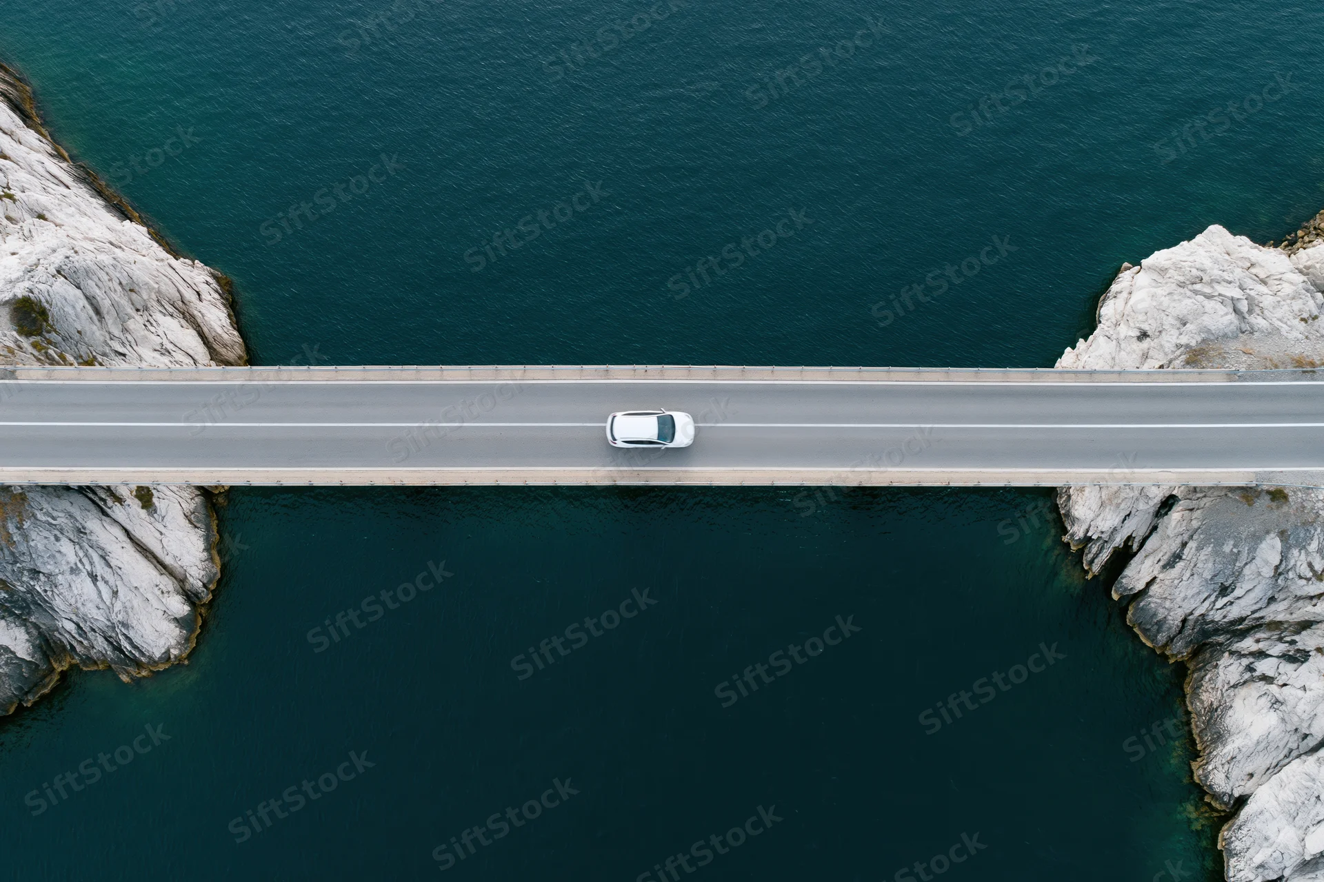 White car driving across narrow concrete bridge over deep blue water between rocky cliffs, aerial top-down view