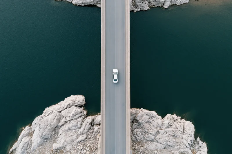 Aerial top-down view of a white car driving on a narrow concrete bridge over dark water between rocky cliffs