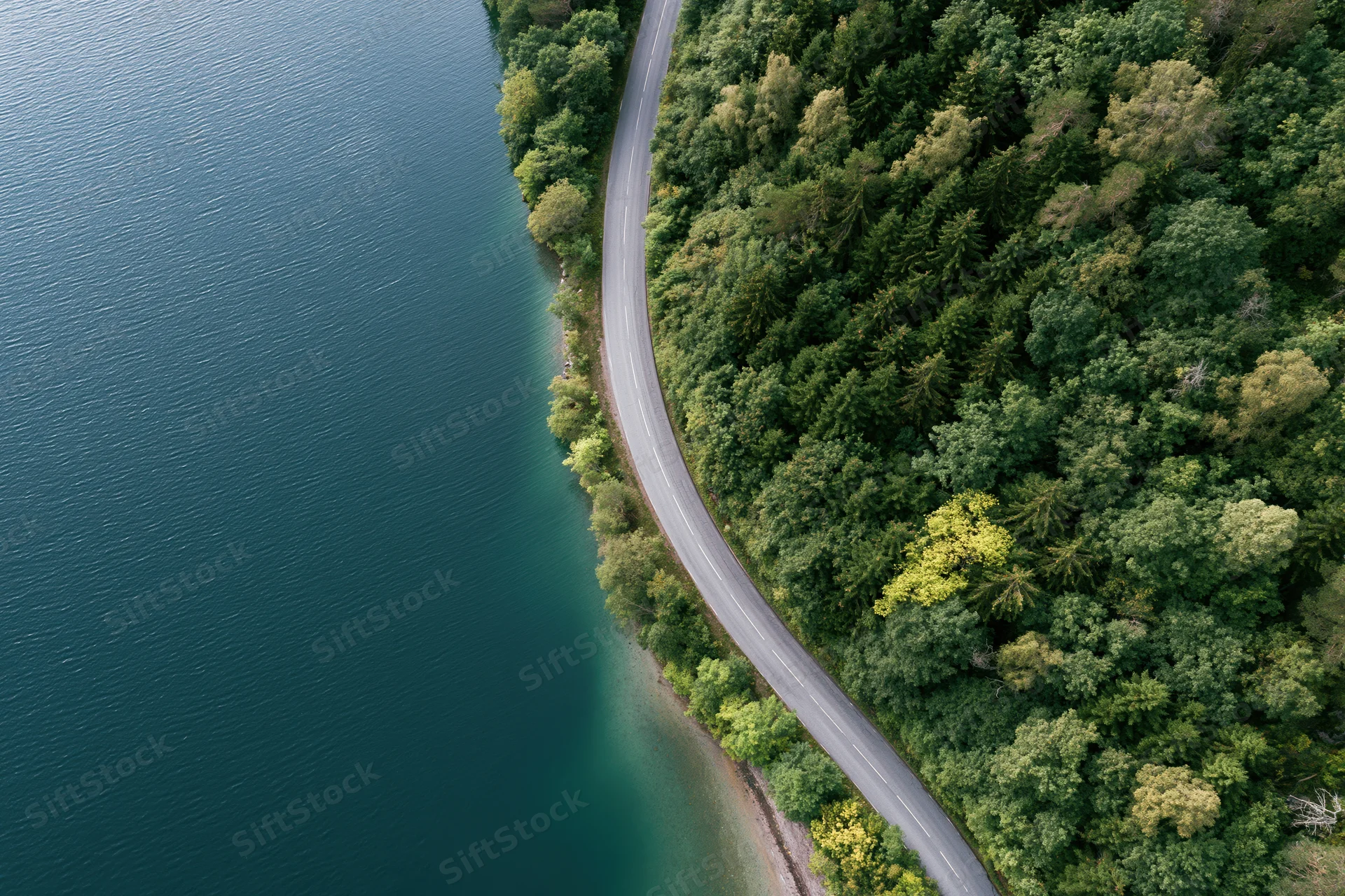 Aerial view of a winding road curving along a forested shoreline beside a calm lake