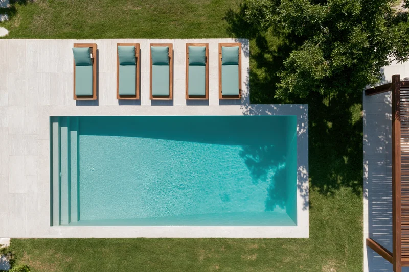 Aerial top-down view of a rectangular outdoor swimming pool with lounge chairs on a tiled deck surrounded by grass and trees