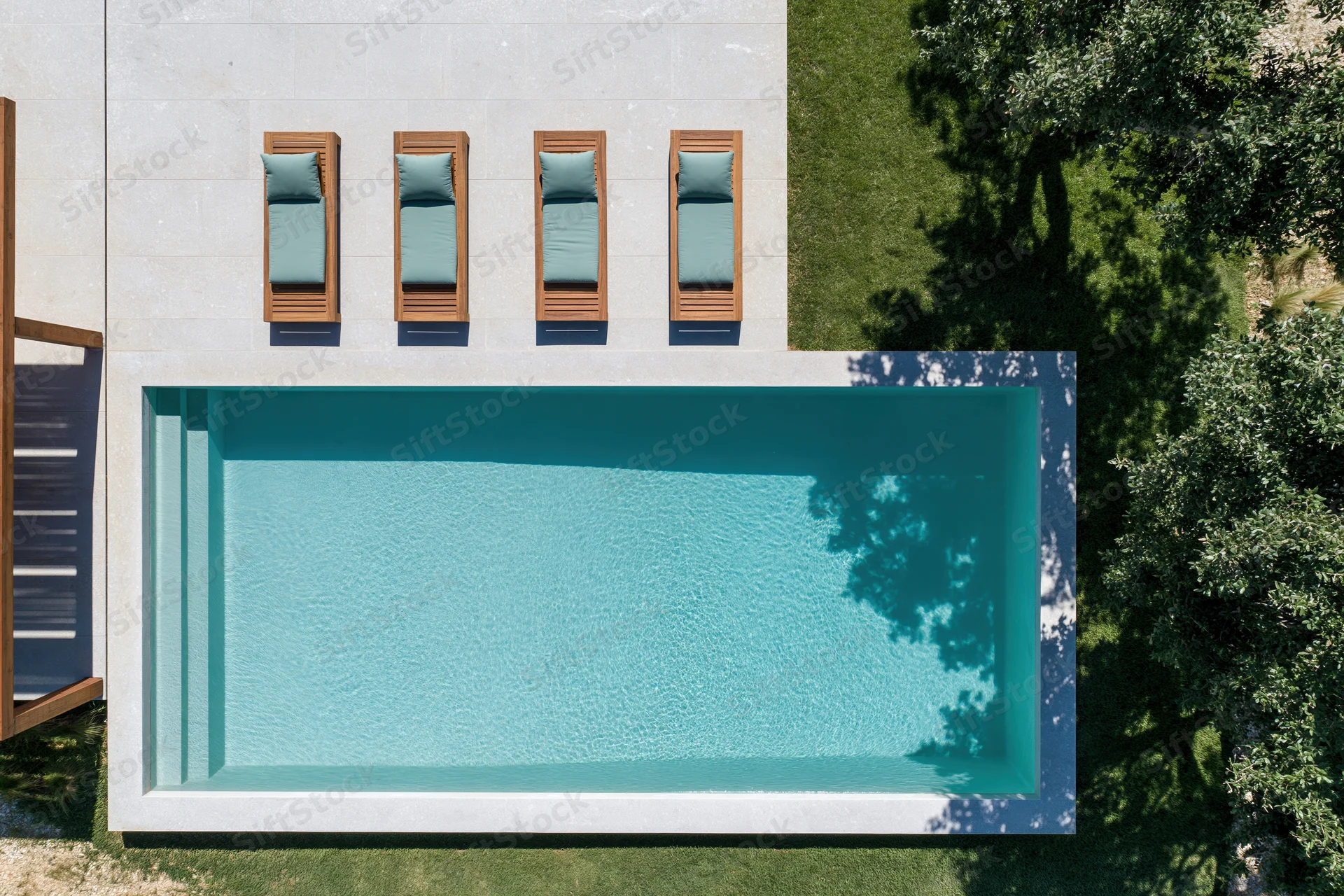 Aerial top-down view of a rectangular outdoor swimming pool with four wooden lounge chairs on a tiled deck beside green grass and trees