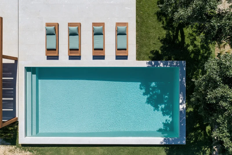Aerial top-down view of a rectangular outdoor swimming pool with four wooden lounge chairs on a tiled deck beside green grass and trees
